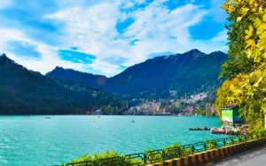 Beautiful view of Naini Lake with blue sky, clouds, and green trees – Nainital tour package highlight.