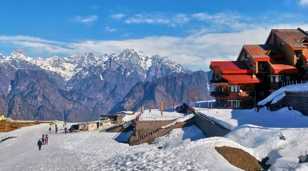 Snow-covered slopes of Auli in winter with pine trees and distant Himalayan peaks under a clear blue sky.