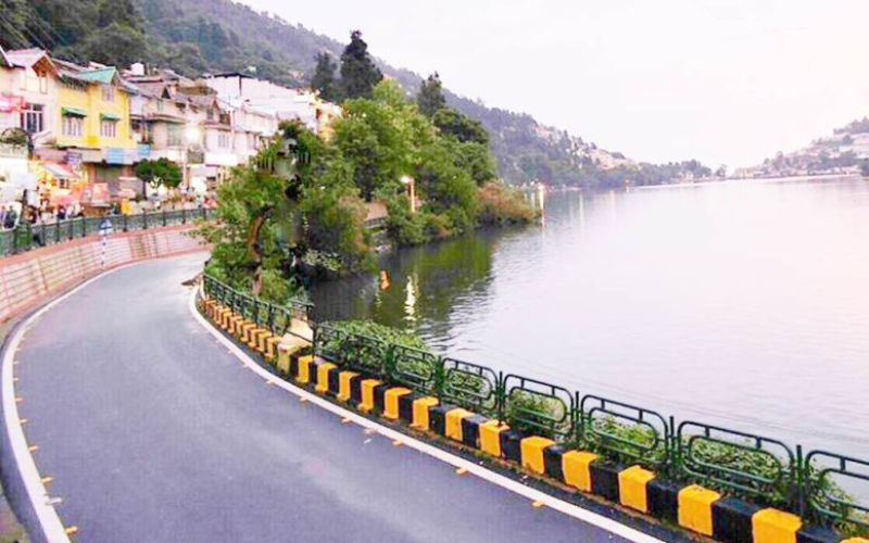 "A scenic view of Naini Lake in Nainital, surrounded by lush green hills, with colorful boats floating on the calm water. Mall Road lines the shore, bustling with visitors and lined with shops and colonial-era buildings. The snow-capped Himalayas can be seen in the distant backdrop under a clear blue sky."