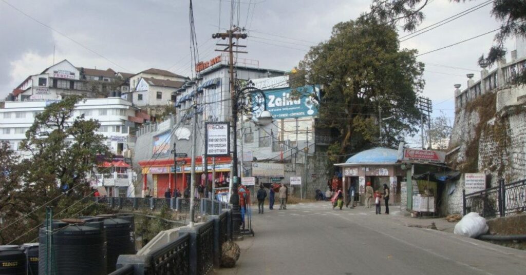 "Mall Road in Mussoorie - a bustling promenade lined with shops, cafes, and colonial-era architecture, surrounded by misty Himalayan hills."
