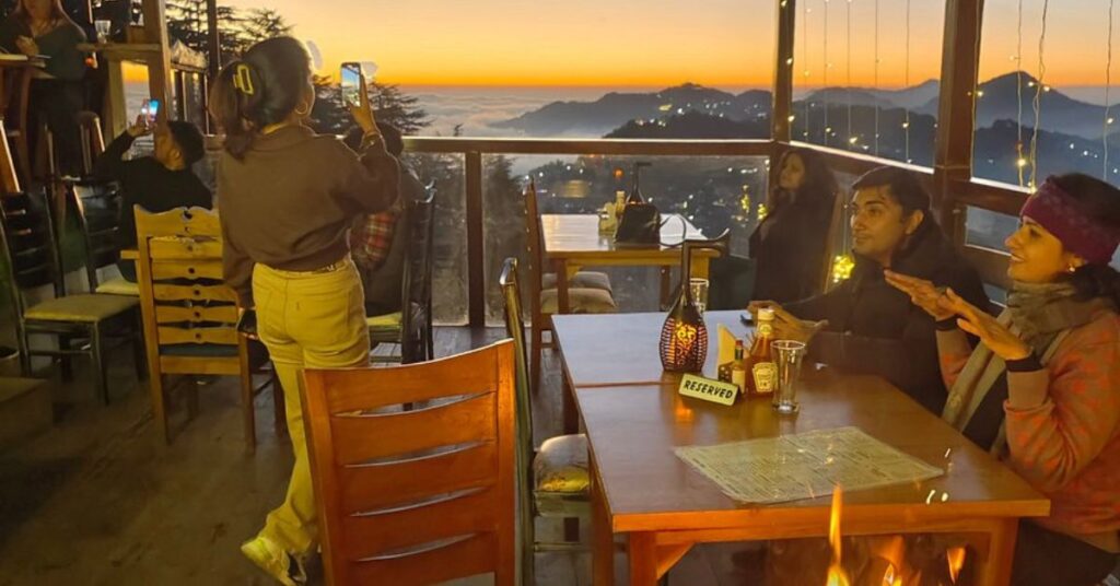 "Tourists savoring hot snacks and live music at a cozy Mussoorie hillside cafΓ©, with misty mountain views and rustic wooden dΓ©cor."