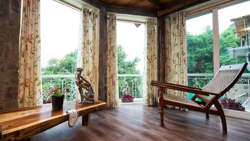 A cozy homestay nestled in the lush green hills of Bhowali, with a charming wooden porch, potted plants, and scenic mountain views. The rustic yet elegant design blends with nature, offering a peaceful retreat.