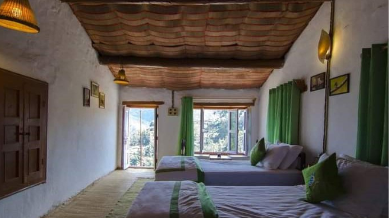 "A traditional Kumaoni stone-and-wood homestay nestled in a vibrant village, surrounded by terraced fields and oak forests. The rustic architecture features carved balconies, with panoramic views of the Himalayan foothills."