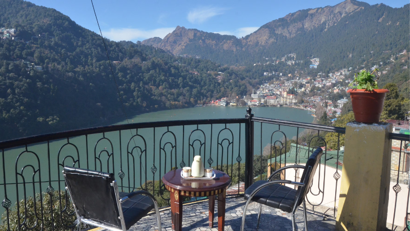 "A serene terrace view from Innisfree Homestay in Tallital, showcasing Naini Lake's shimmering blue waters surrounded by lush green hills, with colorful boats dotting the shoreline and the Himalayan foothills in the distance."